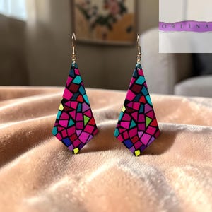Puede incluir: Un par de pendientes colgantes geométricos con diseño de vidriera. Los pendientes presentan un patrón de mosaico de formas rosas, rojas, azules, verdes y amarillas. Cuelgan de ganchos dorados y se exhiben sobre una superficie color melocotón.