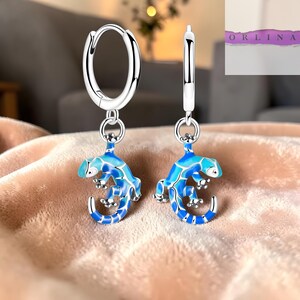 Peut inclure: Boucles d'oreilles créoles en argent avec des breloques en forme de lézard bleu et turquoise. Les lézards présentent un travail d'émail détaillé et sont suspendus aux créoles par de petits connecteurs. Le nom de la marque 'ORLINA' est visible.