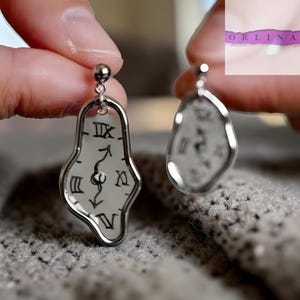 Puede incluir: Pendientes plateados con un diseño único de esfera de reloj derritiéndose. Los pendientes presentan números romanos y manecillas negras, creando un aspecto surrealista y artístico. El diseño recuerda a una pintura famosa.