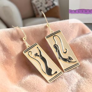 May include: Gold-tone cat earrings. Each earring features a rectangular pendant with a black cat silhouette on a cream background, framed by black and gold lines. The earrings have a hook closure.