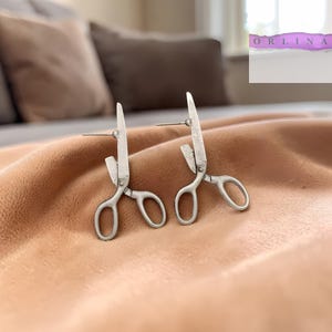 May include: A pair of silver-colored scissor-shaped earrings. The earrings are detailed with a small loop at the end of the handle and a pointed blade. The earrings are displayed on a peach-colored fabric.