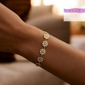 May include: A delicate gold-tone chain bracelet featuring a series of small daisy charms with white petals and yellow centers. The bracelet is linked together in a repeating floral pattern. The text ORLINA is visible in the upper right corner.