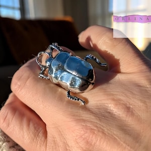 May include: A silver-toned ring featuring a large, three-dimensional beetle design with a smooth, reflective surface. The insect-shaped ornament covers the top of the finger, showcasing detailed legs and a rounded shell. The text ORLINA is visible.