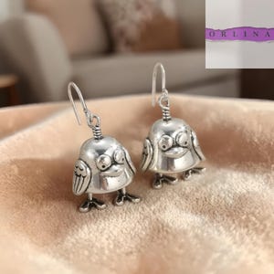 Peut inclure: Une paire de boucles d'oreilles argentées en forme d'oiseau. Chaque boucle d'oreille représente un oiseau avec un corps en forme de cloche, des ailes détaillées et de petits pieds. Les boucles d'oreilles ont une fermeture à crochet.