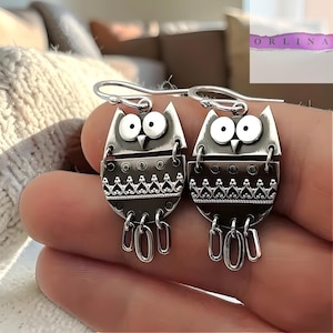 Peut inclure: Boucles d'oreilles argentées en forme de hibou avec un design détaillé. Chaque boucle d'oreille représente un hibou stylisé avec de grands yeux, un motif décoratif et des breloques ovales pendantes. Les boucles d'oreilles sont suspendues à un crochet argenté.