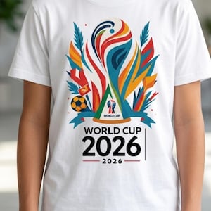 以下が含まれることがあります： 2026年ワールドカップのカラフルなグラフィックデザインが施された白いTシャツ。サッカーボール、赤、青、オレンジ、緑の抽象的な形、そして「WORLD CUP 2026」の文字が含まれています。