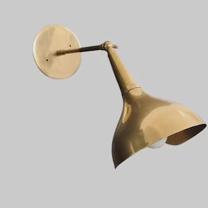 May include: A brass-toned wall sconce with a circular backplate and a curved, cone-shaped shade. The sconce has a long arm connecting the backplate to the shade, and a light bulb is visible inside the shade. The fixture is designed for wall mounting.