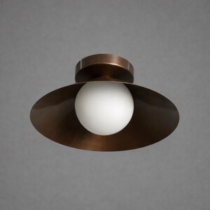 May include: A flush mount ceiling light fixture featuring a wide, circular, dark bronze metal shade with a smooth finish. Centered within the shade is a single, spherical, opaque white glass globe that serves as the light diffuser for the lamp.