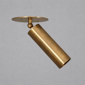 May include: A brass-colored, cylindrical spotlight fixture. The light is mounted on a circular base and has an adjustable ball joint. The cylindrical light is angled downward. The fixture is made of metal and has a smooth finish.