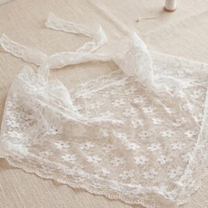 May include: A delicate white lace scarf with a floral pattern and scalloped edges. The scarf has long ties for securing and is made of a sheer, lightweight fabric. A spool of thread and a pin are in the background.