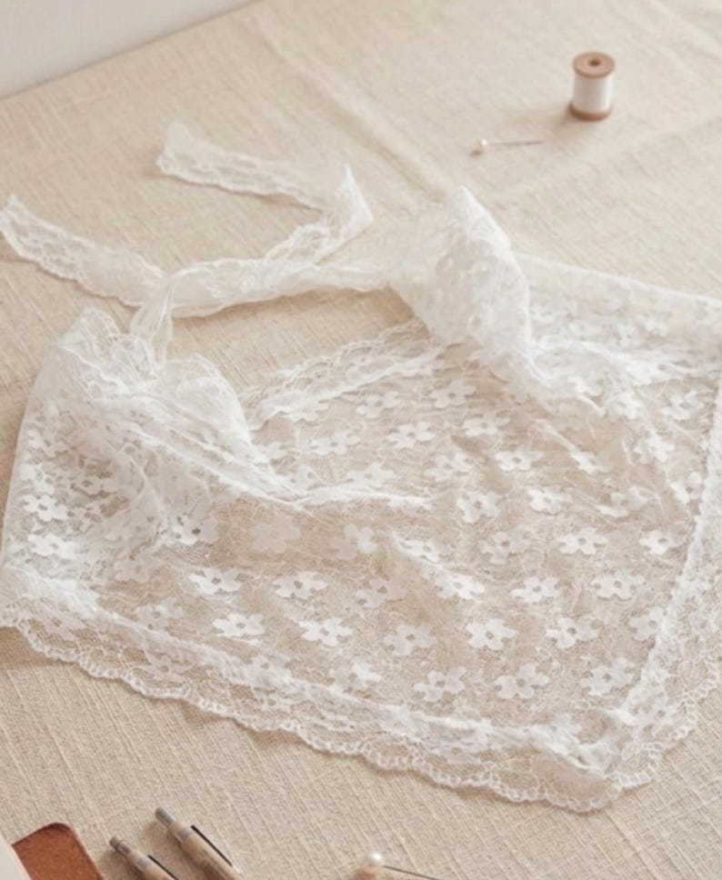 May include: A delicate white lace scarf with a floral pattern and scalloped edges. The scarf has long ties for securing and is made of a sheer, lightweight fabric. A spool of thread and a pin are in the background.
