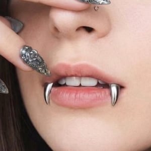Könnte beinhalten: Nahaufnahme eines Gesichts mit silbernen Fangzähnen und kunstvollen, langen, silbernen Nageldesigns. Die Fangzähne werden auf der Unterlippe getragen, und die Nägel haben detaillierte Muster. Ein Nasenpiercing ist ebenfalls sichtbar.