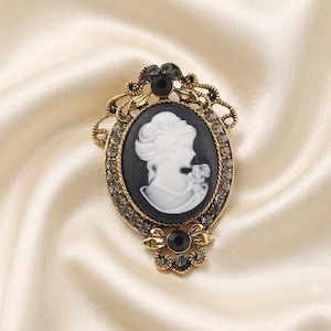 Puede incluir: Un broche ovalado con un camafeo blanco de perfil de una persona sobre fondo negro, engastado en un marco de metal dorado ornamentado. El marco está decorado con pequeños cristales transparentes, dos piedras negras circulares y filigrana.