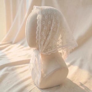 May include: A white lace head covering with a floral pattern and scalloped edges is draped over a mannequin head. The head covering is made of delicate, sheer fabric. The background is a soft, neutral color.