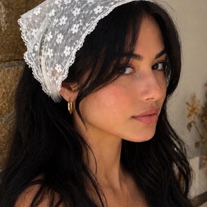May include: A white lace headscarf with a floral pattern. The headscarf is worn on the head. The person is wearing gold hoop earrings and a gold necklace.