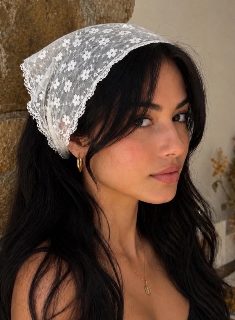 May include: A white lace headscarf with a floral pattern. The headscarf is worn on the head. The person is wearing gold hoop earrings and a gold necklace.