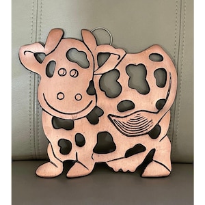 May include: A copper-colored cow-shaped wall hanging with black spots and cut-out details. The cow has a smiling face and a ring for hanging. The design is rustic and decorative, suitable for a kitchen or farmhouse decor.