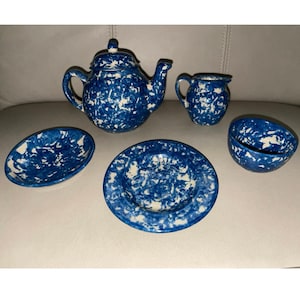 May include: A blue and white ceramic tea set featuring a teapot, creamer, sugar bowl, plate, and an oval dish. The set has a speckled pattern. The teapot has a lid and a curved spout. The creamer has a handle. The plate and oval dish are shallow.