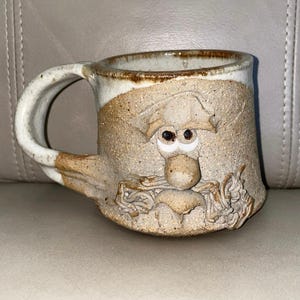 Op de afbeelding: Een handgemaakte keramische mok met een grillig gezichtsontwerp. De mok heeft een bruin en wit kleurenschema met een gebeeldhouwd gezicht, inclusief ogen, een neus en een baard. De handgreep is gebogen en aan de zijkant van de mok bevestigd.