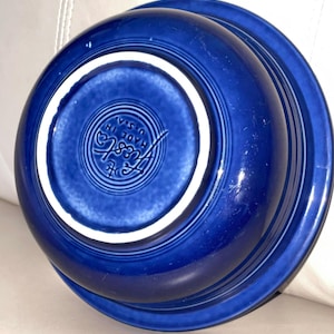 May include: A deep cobalt blue ceramic bowl with a glossy finish. The underside features a circular embossed stamp that reads Fiesta Made in U.S.A. surrounded by concentric rings. The base rim is unglazed white ceramic, contrasting with the blue glaze.