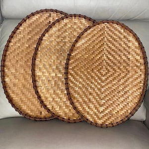 Puede incluir: Tres bandejas redondas de bambú tejido, de color marrón claro natural. Cada bandeja tiene un borde marrón oscuro. Las bandejas están dispuestas superpuestas, destacando su intrincado diseño tejido.