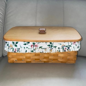 May include: A rectangular woven basket with a wooden lid and handle. The basket has a floral fabric trim with pink, green, and yellow flowers. The basket is made of light brown woven material.