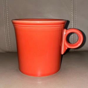 May include: A vibrant orange ceramic mug with a distinctive handle. The mug has a cylindrical body with a slightly flared rim and a circular handle. The exterior is a solid, glossy orange color, and the mug appears to be a classic design.