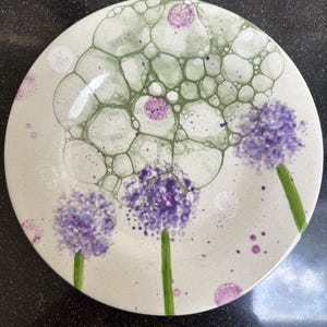 May include: A ceramic plate with a whimsical design. The plate features purple flower-like shapes with green stems and a bubble-like pattern in shades of green and purple. The plate has a white background.