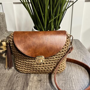 May include: A brown crocheted crossbody bag with a brown leather flap and a gold clasp. The bag has a long brown strap and a brown tassel.