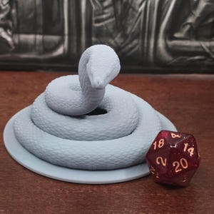 May include: A light blue, coiled snake figurine on a matching round base, with a red and gold twenty-sided die. The snake is the primary focus, with detailed scales. The die is positioned to the right of the snake, on a brown surface.