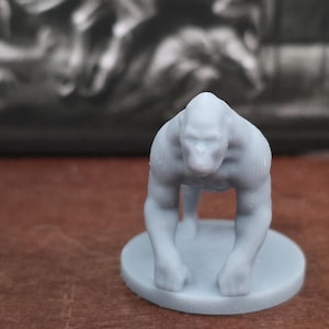 May include: A light gray gorilla figurine in a quadrupedal stance, mounted on a circular base. The gorilla has a muscular build and is facing forward. The background is a blurred image of a dark, textured surface.
