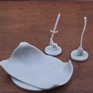 May include: A light gray miniature set featuring a cloak, a sword, and a broom. The cloak is a flat, curved shape, and the sword and broom are on circular bases. The items are likely for tabletop gaming.