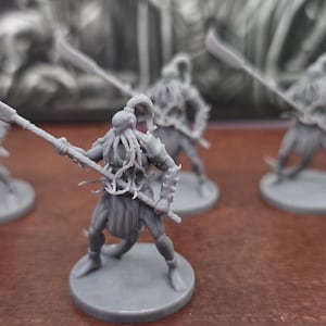 May include: A collection of gray miniature figures of warriors, each holding a spear with a curved blade. The figures are on circular bases, with detailed armor and headgear. The background is blurred.