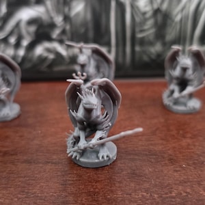 May include: Four gray dragon miniatures with outstretched wings and swords, set on a brown surface. The dragons are detailed with textured scales and stand on circular bases. The background features a blurred, dark relief.