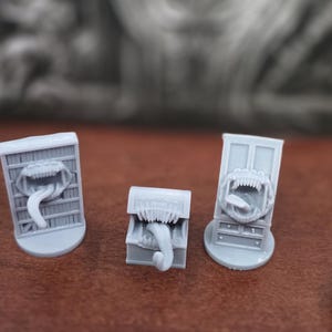 May include: Three gray miniature monster-themed figurines. Each has a mouth with sharp teeth and a long tongue. One is a chest, and the others are framed like doors. The background is blurred.