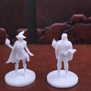 May include: Two white miniature figurines of a wizard and a warrior. The wizard wears a pointed hat and cape, holding a flame. The warrior stands with a cape and a weapon. Both are on circular bases, set against a brown background.