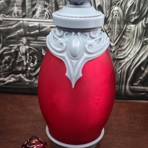 May include: A red, egg-shaped container with a decorative, light gray lid and base. The container has a detailed, ornate design. A red and brown, multi-sided die with white numbers is next to the container.