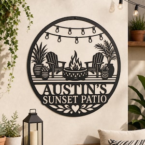 May include: A circular black metal wall sign featuring a cutout design of two chairs, a fire pit, potted plants, and string lights. The text AUSTIN'S SUNSET PATIO is centered below the scene, with a small heart and leaf motif at the bottom.