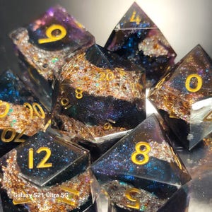 May include: Set of seven polyhedral dice with a galaxy theme. The dice are black with gold numbering and filled with gold glitter, blue and purple swirls. The dice are for tabletop games.