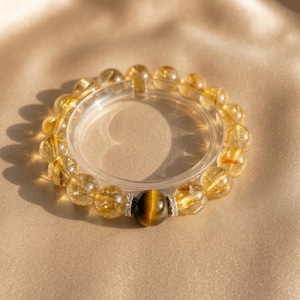 May include: A golden-yellow beaded bracelet with a large tiger's eye bead and clear spacer. The bracelet is resting on a satin surface. The beads have a translucent quality, and the tiger's eye bead has a brown and yellow pattern.