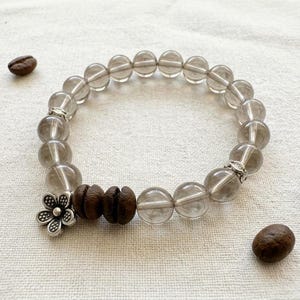 Könnte beinhalten: Ein Armband mit Rauchquarzperlen, Holzperlen und einem silbernen Blumenanhänger. Kaffeebohnen sind um das Armband herum verstreut. Das Armband liegt auf einer weißen Oberfläche.