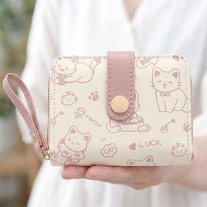 Peut inclure: Un portefeuille crème avec une sangle rose et une fermeture à bouton doré. Le portefeuille présente un motif répété de chats de dessins animés, d'empreintes de pattes et les mots "Meow", "Cat" et "Luck". Le portefeuille a une fermeture éclair et une dragonne.