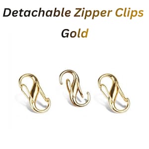 Puede incluir: Tres clips desmontables para cremallera de metal dorado con un acabado liso y pulido. Cada clip cuenta con un mecanismo de cierre de resorte de doble extremo. El texto "Detachable Zipper Clips Gold" aparece en la parte superior.