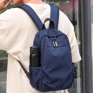 May include: A navy blue backpack with adjustable shoulder straps, a top handle, and a front zippered compartment. The bag features a side pocket containing a matte black cylindrical water bottle with a secure lid and a drawstring closure.