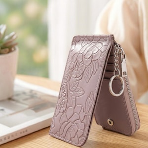 May include: A mauve key case featuring an embossed floral pattern on the exterior. The accessory includes a silver-toned metal chain with a circular key ring attached to the side, a zipper closure, and a snap button fastener on the interior flap.