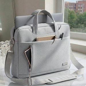 Può includere: Una borsa per computer portatile grigio chiaro dotata di uno scomparto principale con un laptop all'interno, due manici superiori e una tracolla rimovibile. La parte anteriore presenta una tasca orizzontale con un taccuino e una tasca verticale.