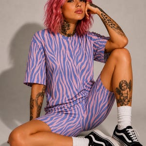 May include: A two-piece set consisting of a short-sleeved t-shirt and matching cycling shorts, both featuring a repeating zebra stripe pattern in light pink and periwinkle blue. The t-shirt has a crew neckline and the shorts are form-fitting.