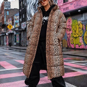 Pode incluir: Um casaco comprido acolchoado com padrão de leopardo castanho e preto, usado sobre uma camisola preta com capuz e calças pretas com costuras brancas. O conjunto inclui óculos de sol retangulares e sapatilhas pretas com plataforma.