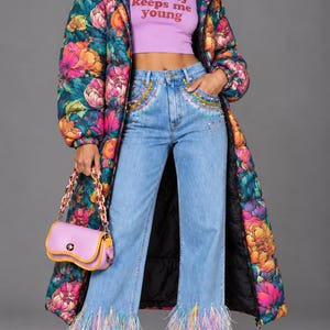 Può includere: Un lungo piumino floreale con base scura e fiori vivaci rosa, arancioni e blu, un top corto color lavanda con la scritta insanity keeps me young, jeans azzurri con tasche decorate da perline e orli piumati, e una borsa a spalla color lavanda.