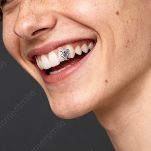 May include: A close-up of a person smiling, showcasing a tooth gem. The gem is silver-colored, featuring a design with a purple and blue gemstone. The person's teeth are white, and the lips are a natural pink. The background is a solid gray.